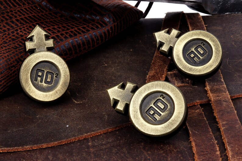 May include: Three gold-colored metal tokens with a symbol and the letters "AD" with a superscript "2" on each. The tokens are on a brown leather surface.