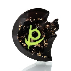 May include: Black resin pendant with a green and gold symbol, resembling a crescent moon with a circle inside. The pendant is embedded with gold flakes.