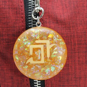 May include: A round, gold-colored keychain with a white symbol embedded in the resin. The keychain is filled with iridescent glitter.