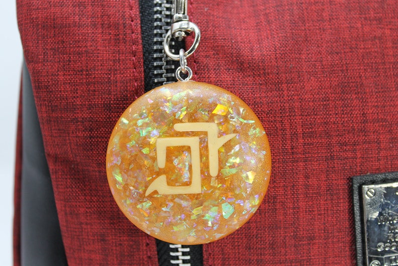 May include: A round, gold-colored keychain with a white symbol embedded in the resin. The keychain is filled with iridescent glitter.