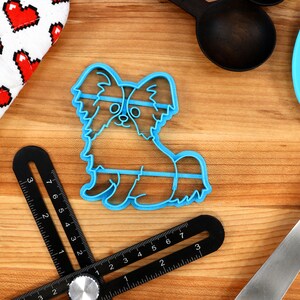 May include: Blue plastic cookie cutter shaped like a papillon dog. The cutter is on a wooden surface with other baking tools.