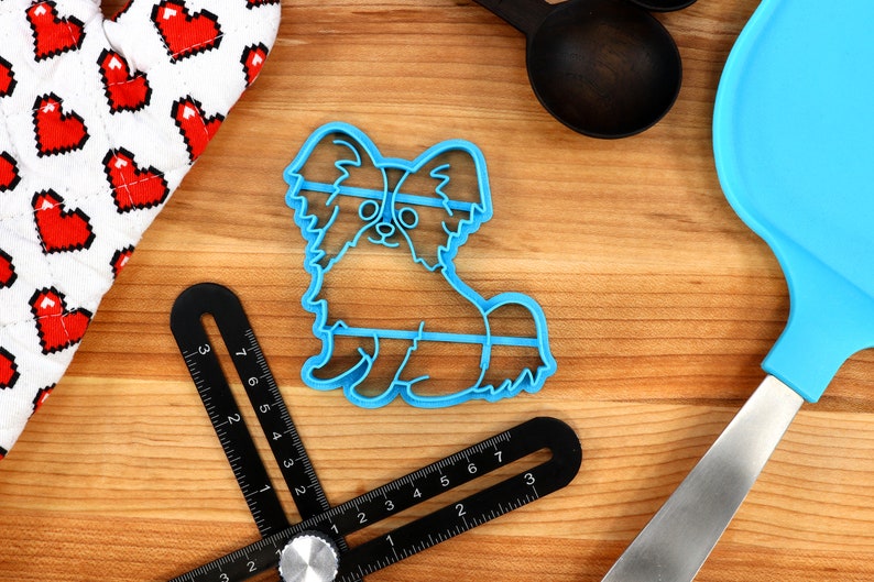 May include: Blue plastic cookie cutter shaped like a papillon dog. The cutter is on a wooden surface with other baking tools.
