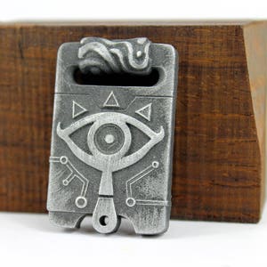 May include: A silver-coloured metal pendant featuring a stylised eye design and geometric patterns. The pendant has a loop at the top for attaching to a chain or cord.