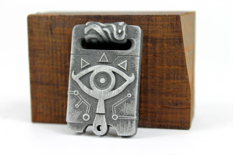 May include: A silver-coloured metal pendant featuring a stylised eye design and geometric patterns. The pendant has a loop at the top for attaching to a chain or cord.
