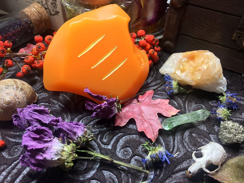 May include: Orange candle with three carved lines on a dark fabric background with dried flowers, a seashell, a small skull, and a green crystal.