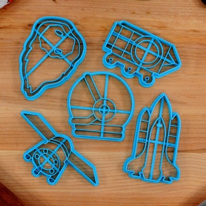May include: A set of six blue plastic cookie cutters shaped like space-themed objects, including a rocket, a satellite, a space helmet, a spaceship, and two other space-related shapes. The cutters are on a wooden surface.