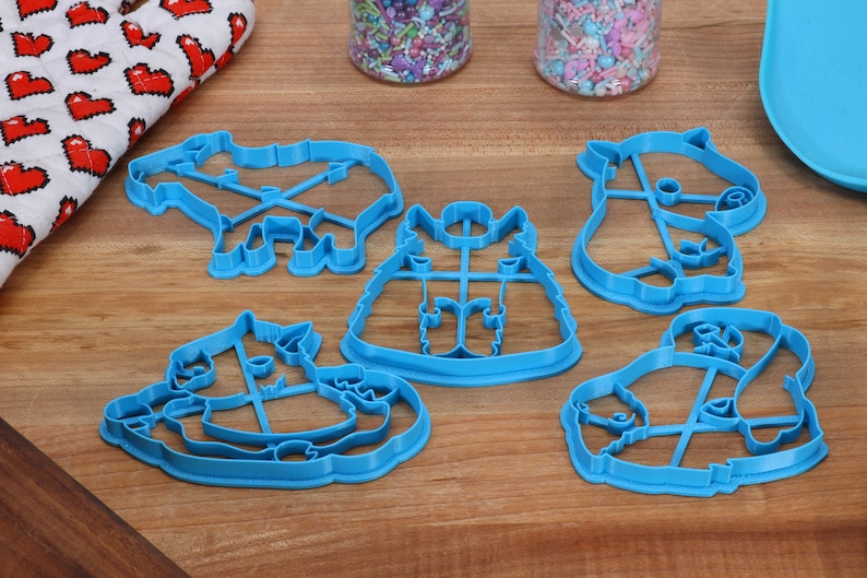 May include: Five blue plastic cookie cutters shaped like animals. The cutters are on a wooden surface. The cutters are for making cookies in the shapes of a unicorn, a cat, a panda, a llama, and a reindeer.