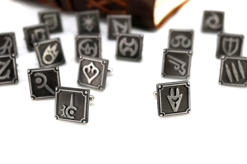 May include: A set of silver cufflinks featuring various symbols, including a sword, a shield, and a crescent moon. The cufflinks are scattered on a white surface.