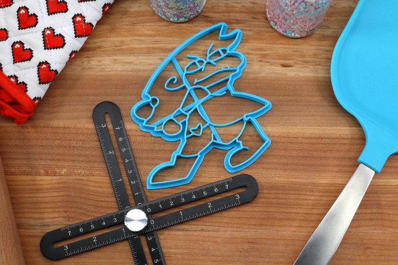 Puede incluir: Un cortador de galletas de pl&aacute;stico azul con forma de personaje de dibujos animados con un sombrero y una espada. El cortador de galletas est&aacute; sobre una superficie de madera con una regla y una esp&aacute;tula.