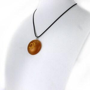 Puede incluir: Un collar con un colgante redondo de color dorado en un cord&oacute;n negro. El colgante tiene un s&iacute;mbolo dorado.