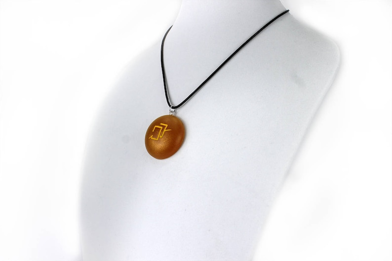 Puede incluir: Un collar con un colgante redondo de color dorado en un cord&oacute;n negro. El colgante tiene un s&iacute;mbolo dorado.