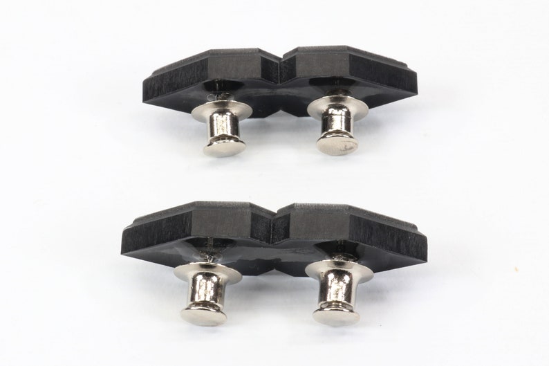 May include: Two black, geometric-shaped push pins with silver-colored metal posts and heads. The push pins are arranged in two sets of two, with a white background.