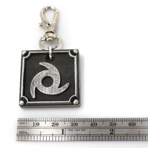 以下が含まれることがあります： 銀色の金属製のキーホルダーで、黒い正方形のチャームに銀色の渦巻き模様が施されています。チャームは約3.5cmの幅です。