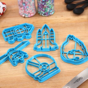 May include: A set of five blue plastic cookie cutters shaped like a rocket, a house, a spaceship, a helmet, and a dog. The cutters are on a wooden surface.