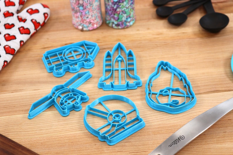 May include: A set of five blue plastic cookie cutters shaped like a rocket, a house, a spaceship, a helmet, and a dog. The cutters are on a wooden surface.