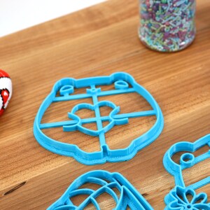 May include: A set of blue plastic cookie cutters shaped like a bear, a frog, a flower, and a heart. The cutters are on a wooden surface.
