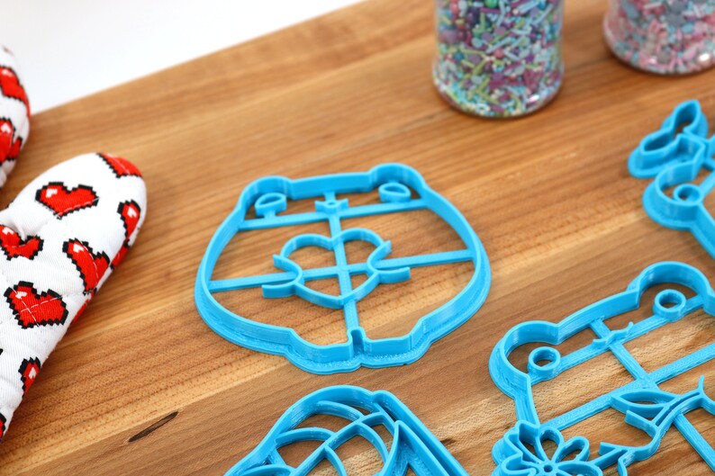 May include: A set of blue plastic cookie cutters shaped like a bear, a frog, a flower, and a heart. The cutters are on a wooden surface.