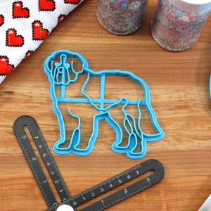 May include: A light blue plastic cookie cutter in the shape of a dog, with a detailed outline. The cutter is on a wooden surface, alongside a blue spatula, a heart-patterned oven mitt, and jars of sprinkles. A black measuring tool is also present.