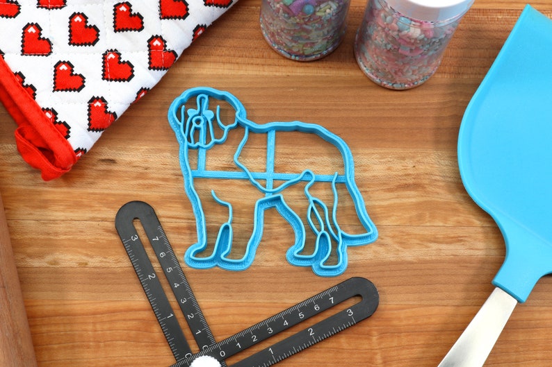 May include: A light blue plastic cookie cutter in the shape of a dog, with a detailed outline. The cutter is on a wooden surface, alongside a blue spatula, a heart-patterned oven mitt, and jars of sprinkles. A black measuring tool is also present.