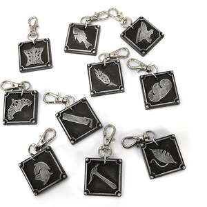 May include: Ten silver keychains with different symbols on black square tags. The symbols include a crown, a fish, an anvil, a wheat stalk, a leaf, a saw, a helmet, a pickaxe, and two more leaves.