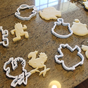 May include: A collection of cookie cutters and shaped cookies on a countertop. The cutters are white plastic in various shapes, including a cactus, a bird, and a character. The cookies are light brown and baked.
