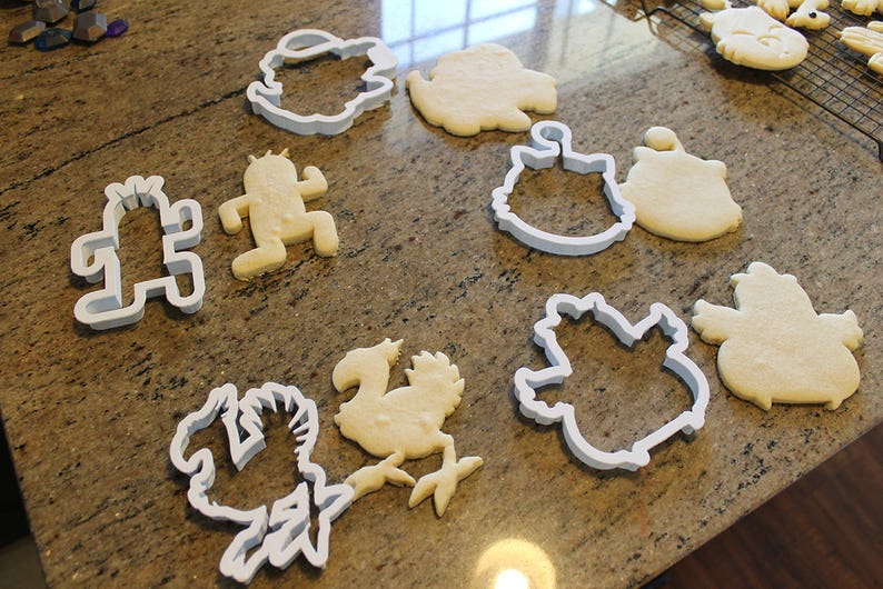 May include: A collection of cookie cutters and shaped cookies on a countertop. The cutters are white plastic in various shapes, including a cactus, a bird, and a character. The cookies are light brown and baked.