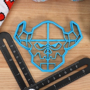 May include: A blue plastic cookie cutter shaped like a geometric skull with horns. The cutter is on a wooden surface with a ruler.