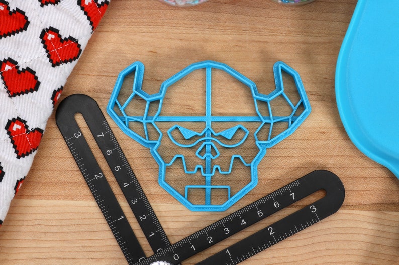 May include: A blue plastic cookie cutter shaped like a geometric skull with horns. The cutter is on a wooden surface with a ruler.