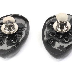 May include: Two black and silver metal pin backs with a textured surface and a silver metal pin. The pin backs are oval shaped.