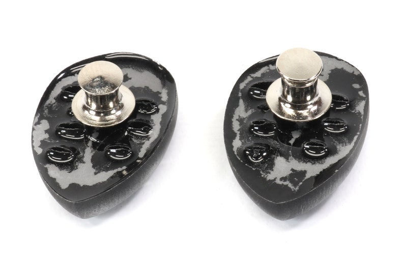 May include: Two black and silver metal pin backs with a textured surface and a silver metal pin. The pin backs are oval shaped.