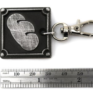 May include: A silver-colored keychain with a black square charm featuring a silver-colored 3-leaf clover design. The keychain has a silver-colored metal clasp.