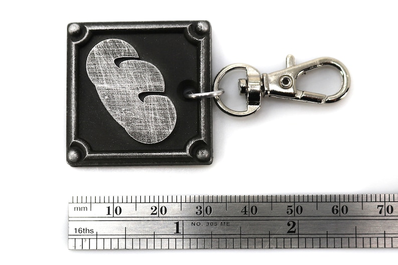 May include: A silver-colored keychain with a black square charm featuring a silver-colored 3-leaf clover design. The keychain has a silver-colored metal clasp.
