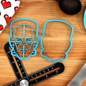May include: Two blue plastic cookie cutters, one shaped like a Frankenstein's monster face and the other a simple outline of a head. The cutters are on a wooden surface with a ruler and a blue spatula.