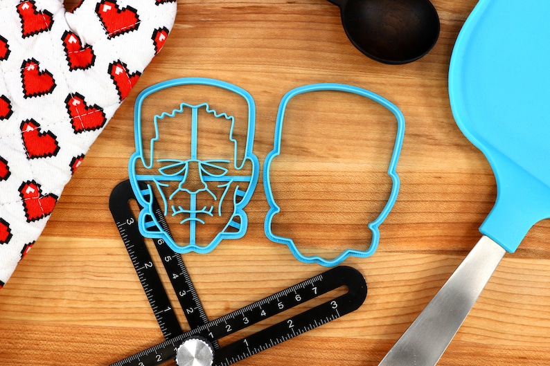 May include: Two blue plastic cookie cutters, one shaped like a Frankenstein's monster face and the other a simple outline of a head. The cutters are on a wooden surface with a ruler and a blue spatula.