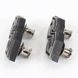 May include: Two geometric-shaped, black and gray cufflinks with silver-colored posts. The cufflinks have a textured surface and are set against a white background. The design is modern and angular.