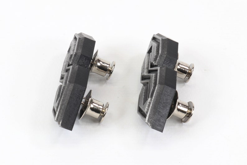 May include: Two geometric-shaped, black and gray cufflinks with silver-colored posts. The cufflinks have a textured surface and are set against a white background. The design is modern and angular.