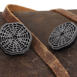 May include: Two silver-colored metal octagonal-shaped decorative pieces with a geometric pattern on a brown leather surface.