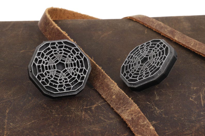 May include: Two silver-colored metal octagonal-shaped decorative pieces with a geometric pattern on a brown leather surface.