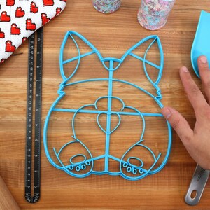 May include: A blue plastic cookie cutter shaped like a dog's rear end. The cutter has a heart-shaped cutout in the center and a detailed outline of the dog's legs and tail.