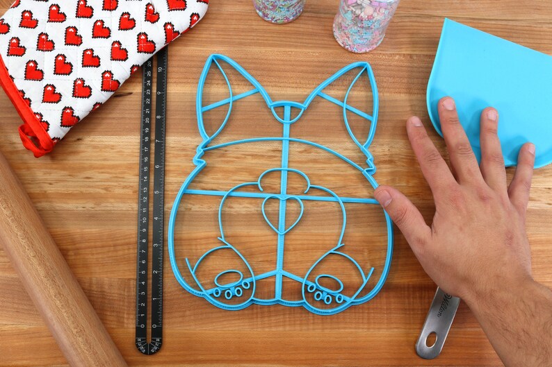 May include: A blue plastic cookie cutter shaped like a dog's rear end. The cutter has a heart-shaped cutout in the center and a detailed outline of the dog's legs and tail.