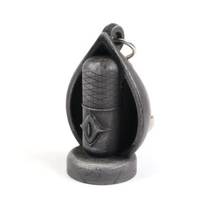 May include: A black metal keychain shaped like a teardrop with a smaller black metal cylinder inside. The cylinder has a diamond-shaped design and a silver ring attached to the top.