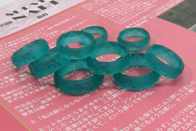 May include: A close-up of nine teal-colored plastic rings with numbers printed on them. The rings are stacked on top of each other and are on a pink surface.