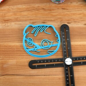 May include: A blue plastic cookie cutter shaped like a cat's face. The cutter is about 2 inches wide and 2 inches tall.