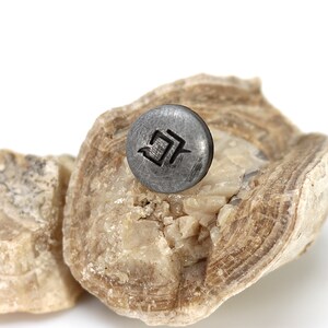 May include: A small, round, dark gray metal object with a geometric design is displayed on a light brown, textured rock formation. The object is centered on the rock, with a white background.