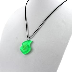 May include: A green pendant necklace with a gold lyre symbol. The pendant is shaped like a leaf and hangs from a black cord.