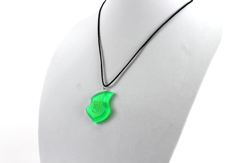 May include: A green pendant necklace with a gold lyre symbol. The pendant is shaped like a leaf and hangs from a black cord.