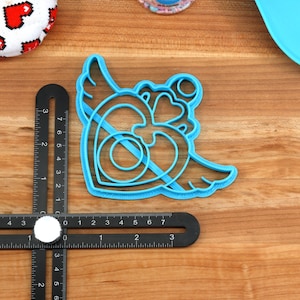 May include: A blue plastic cookie cutter shaped like a heart with wings. The heart has a double outline and a smaller heart inside.