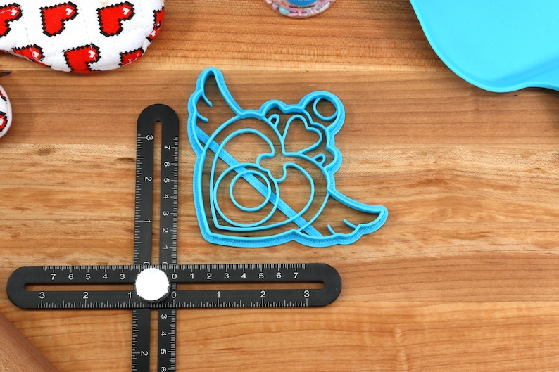 May include: A blue plastic cookie cutter shaped like a heart with wings. The heart has a double outline and a smaller heart inside.