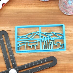May include: A blue plastic cookie cutter shaped like a city skyline with mountains in the background.