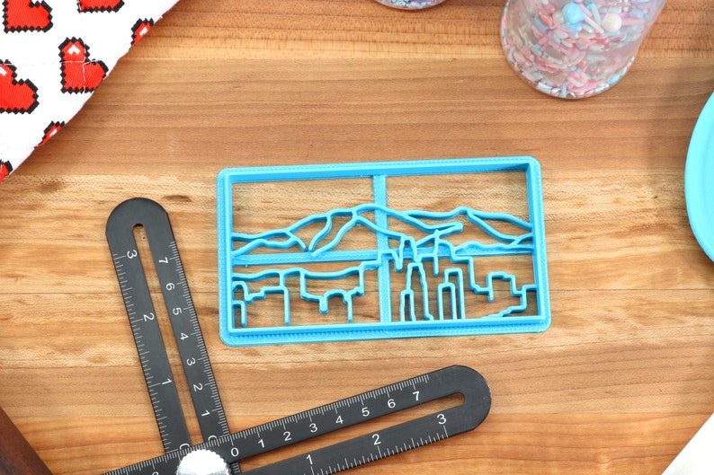 May include: A blue plastic cookie cutter shaped like a city skyline with mountains in the background.